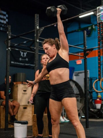 woman in gym standing with one arm extended overhead with a dumbbell in her hand