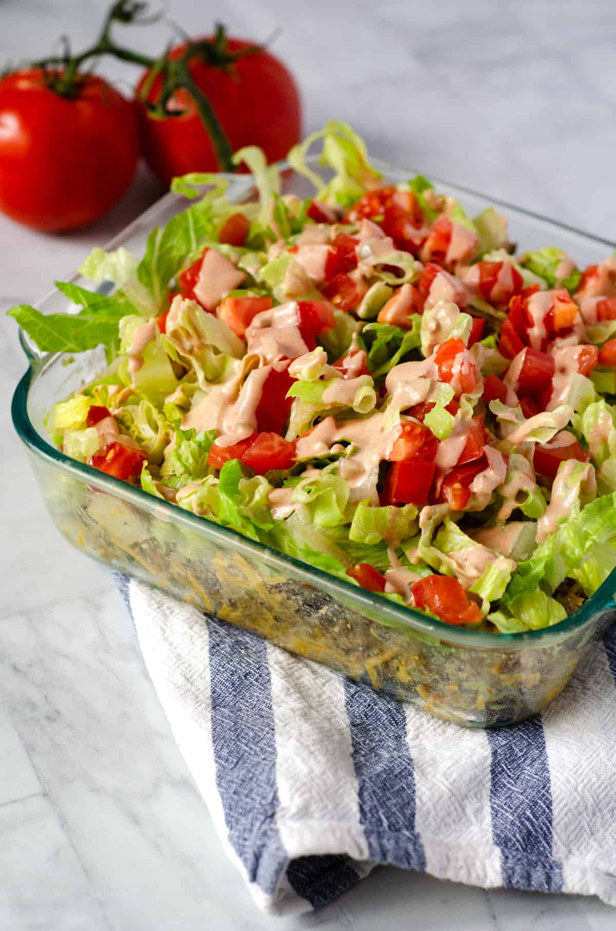 square pan of venison burger casserole topped with lettuce and tomato in a square pan with tomatoes on the vine in the background