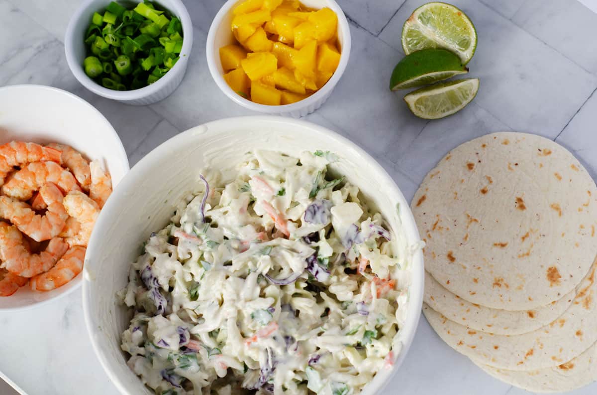 overhead view of bowls of shrimp, coleslaw, mango, green onions, lime slices and some tortillas on a table